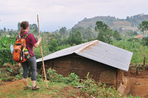 a woman with a backpack standing with a walking stick near a humble house 
