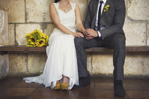 Bride with sunflower bouquet sitting on wooden bench holding hands with groom in chapel.