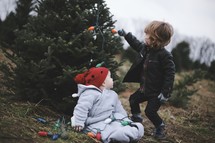 toddler and infant in a Christmas tree lot with Christmas lights 