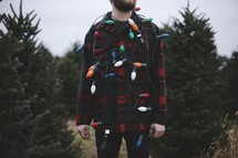 a man in a plaid shirt with Christmas lights standing in a Christmas tree lot 