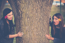 Two girls looking around a tree at each other.