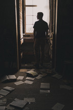 man standing in a window and torn books and pages on the floor 