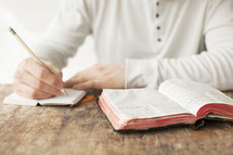 Writing in a journal with a pencil, on a wooden table with an open Bible.