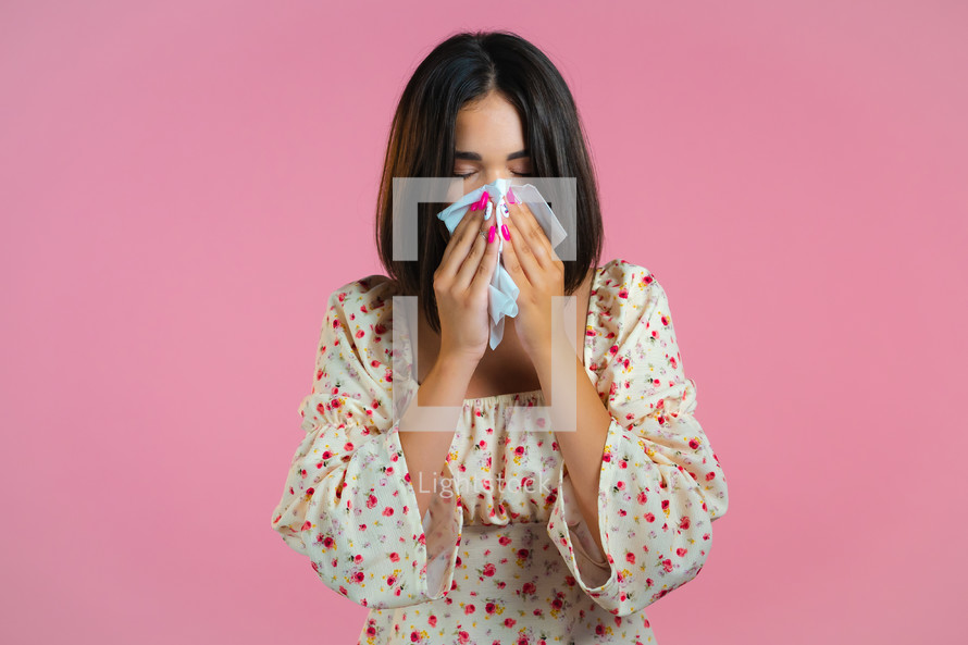 Woman sneezes into tissue. Isolated girl on pink studio background. Lady is sick, has a cold or allergic reaction. Coronavirus, epidemic 2024, illness concept.