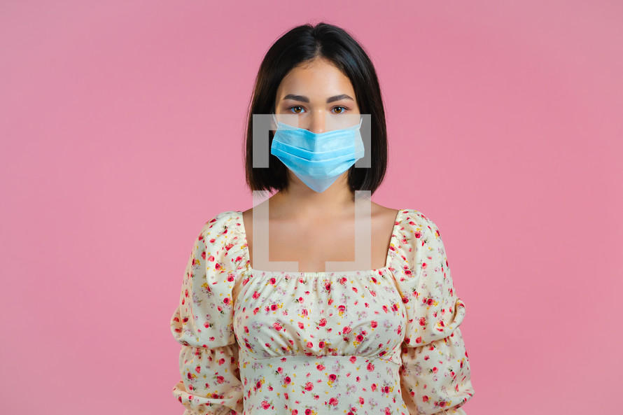 Young pretty girl in face medical mask during coronavirus pandemic. Portrait of girl in floral dress on pink background. Protection with respirator against COVID-19 outbreak.