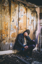 A woman sits in an abandoned house with her face in her hand.
