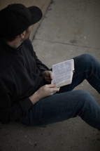 a man sitting on a sidewalk reading a Bible 