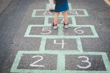 A young girl standing on a hopscotch board with a stuffed animal. 