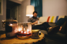 A burning candle and a man reading a Bible on a couch in the distance. 