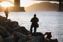 Fisherman on a rocky shore near a bridge.