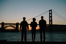 Silhouette of three people looking at a suspension bridge.