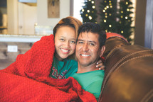 a couple snuggling under a blanket sitting on a couch at Christmas 