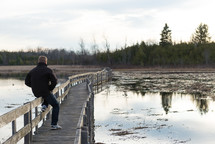 Man sitting on the rail of a wooden bridge over a lake.