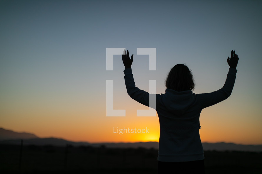 a woman looking out into a field at sunset with raised hands 