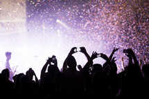 Silhouette of audience at a concert with raised smartphones and confetti in the air.