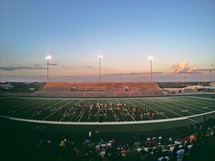 Panoramic view of lighted football field from sideline at dusk.