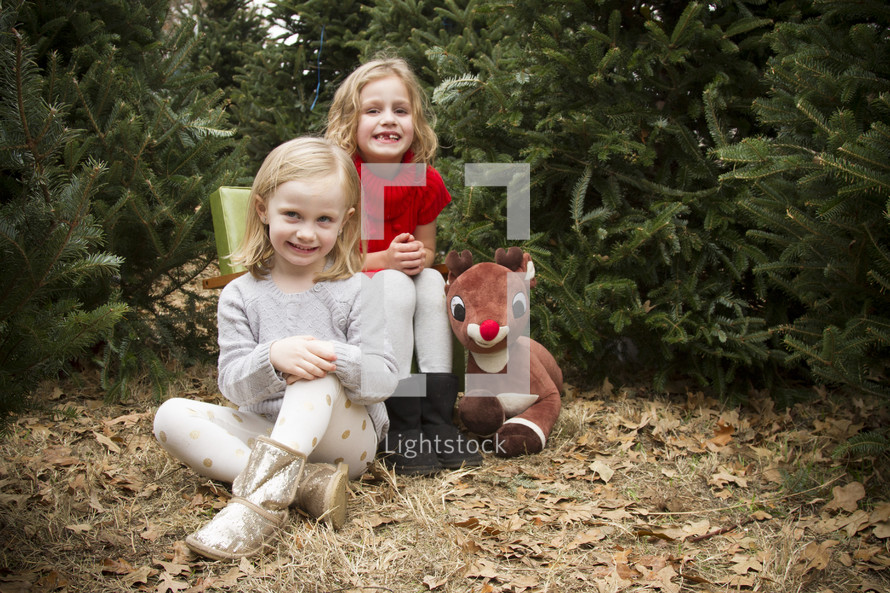 Christmas photos of two sisters and a Rudolph stuffed animal in a Christmas tree lot 