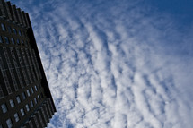 Ground view of side of tall building with cloud formations and blue sky in the background.