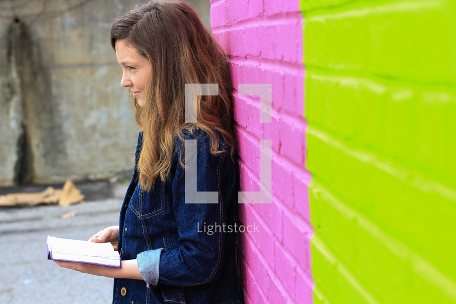 a teen girl in a jean jacket and striped dress reading a Bible in front of a colorful wall 