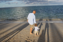 a father walking holding hands on a beach with his toddler son 