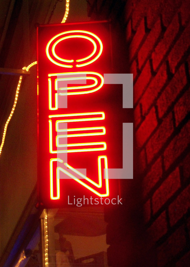 A Neon Open sign lights up the night in neon orange surrounded by Christmas lights illuminating a brick exterior building in a downtown retail area telling passer-bys that the store is open for business and welcoming the public during the shopping retain season.. 