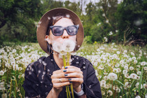 Happy woman blowing on dandelion in park. Girl in hat and sunglasses. Wishing, joy concept.