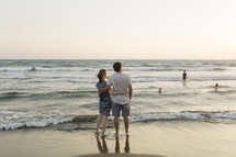 Husband and wife holding each other on a beach while looking at kids in ocean.