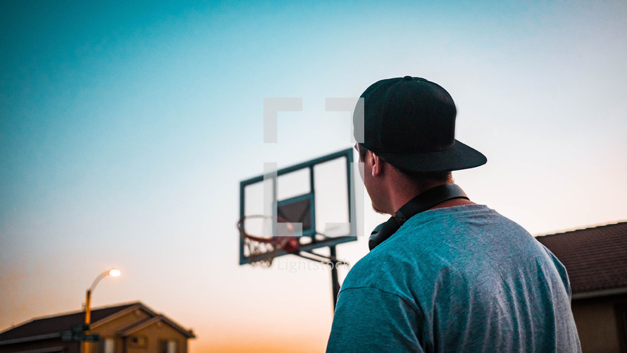 a man outdoors with headphones at sunset looking up at a basketball hoop 