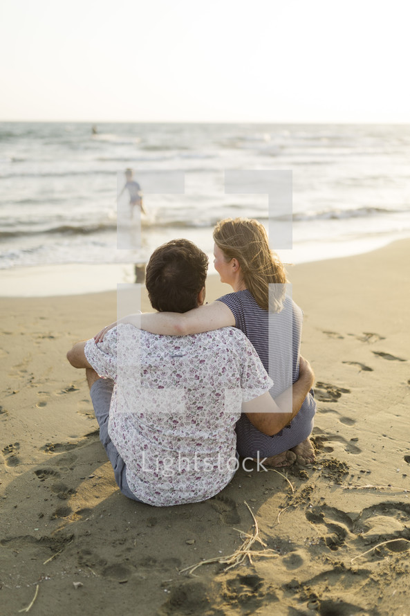 Couple holding each other on the beach while child plays in the ocean.