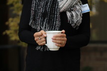 a woman standing outdoors in a scarf holding a mug 