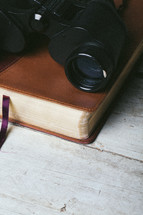 Binoculars on closed Bible laying on top of wooden table.