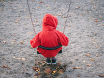 A child in a red hoodie on a sand-covered playground swing looking lonely into the distance