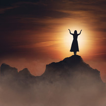 Silhouette of woman with arns raised standing on a mountain top with an orange sunset in the background.