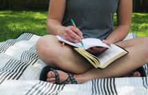 a girl sitting on a blanket in the grass reading a Bible and writing in a journal 