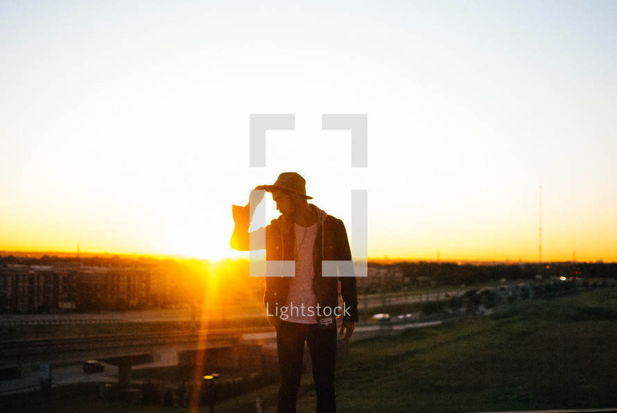 man touching his hat standing outdoors at sunset 