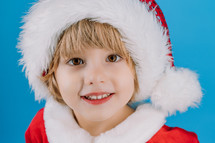 Portrait of adorable boy in Santa Claus hat on blue studio background. Christmas celebration. Happy childhood, kid, lovely son. High quality photo