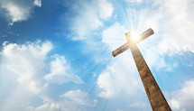 A wooden cross with bright sun and clouds in the background.
