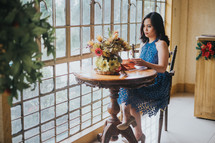 a young woman sitting in front of a window at a table alone with a vase of flowers 