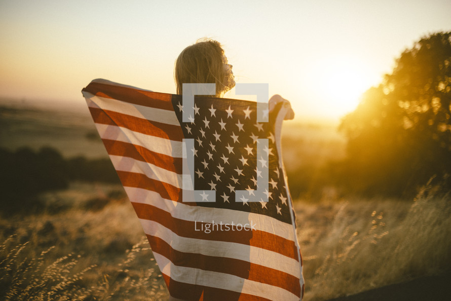 a woman holding an American flag standing in a field at sunset 