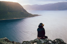 A person sitting on a high cliff overlooking a body of water surrounded by mountains.