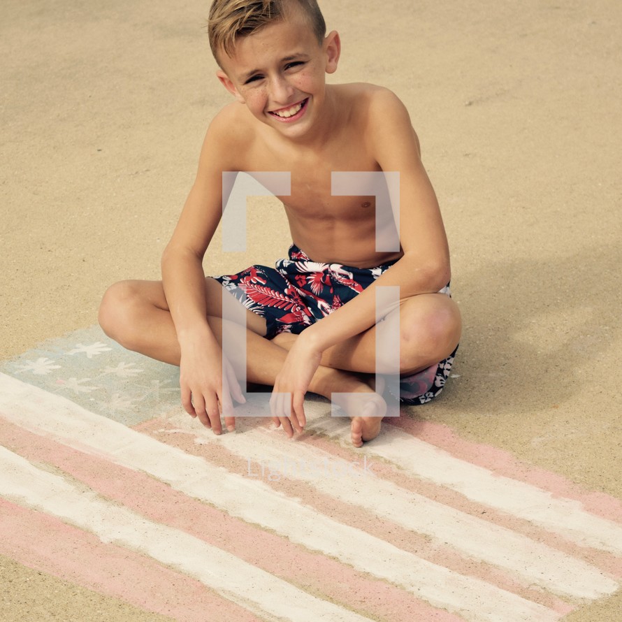 a boy child in a bathing suit sitting on an American flag in sidewalk chalk 