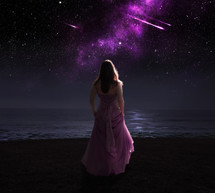 A woman on a beach at night looking at a shooting star. 