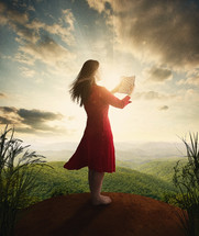 Woman standing on a hill reading the Bible with rays of light beaming from the clouds.