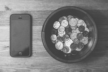 Coins in a bowl and a cell phone on a wooden table.