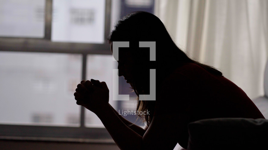Silhouete of hopeful young woman clasping hands in prayer asking for blessing. Religion and faith concept
