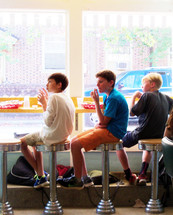 Soda Fountain Kids - A group of three young elementary school boys sit and relax together in a local soda fountain after school eating French fries, hamburgers and soda on a sunny day sitting on round stools in front of a large picture window in a local downtown soda shop restaurant. 