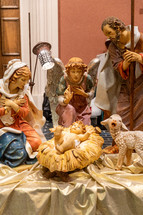 Statues of Mary, Joseph, and an angel facing baby Jesus in the manger; a little lamb stands on the side