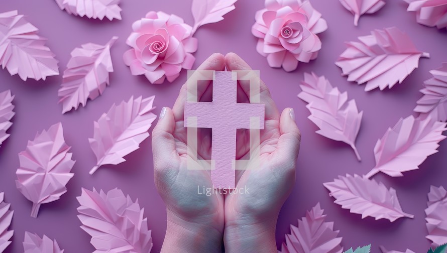 Female hands holding a pink christian cross surrounded by pink paper flowers.