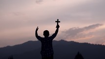 male holding a cross on a mountaintop at sunrise 