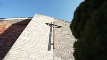 A wooden cross on a brick wall with a green bush in front of a church with bright shining light.